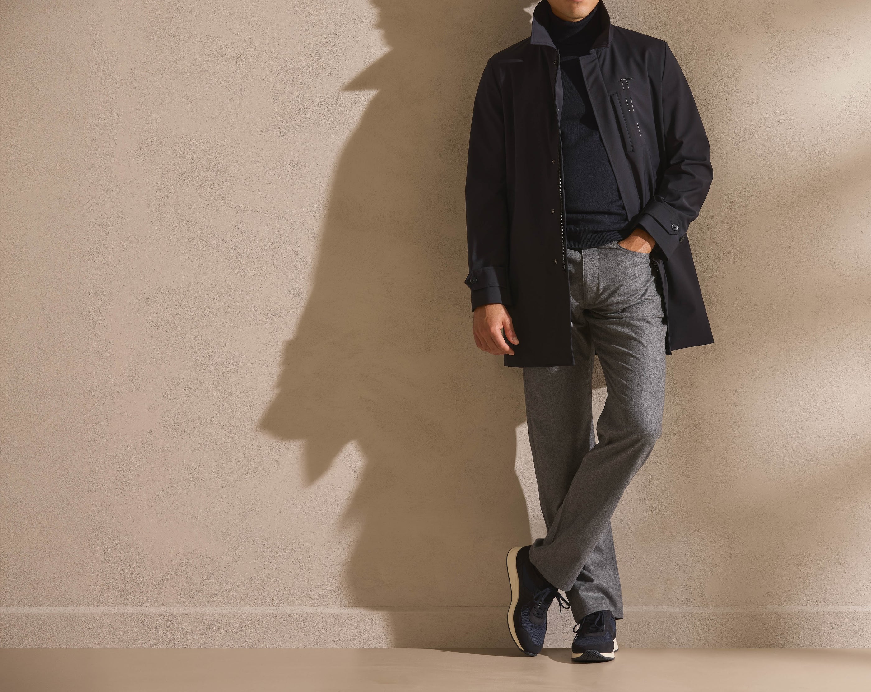 A navy technical coat over fine knitwear, with grey trousers and runners — utility, refined.