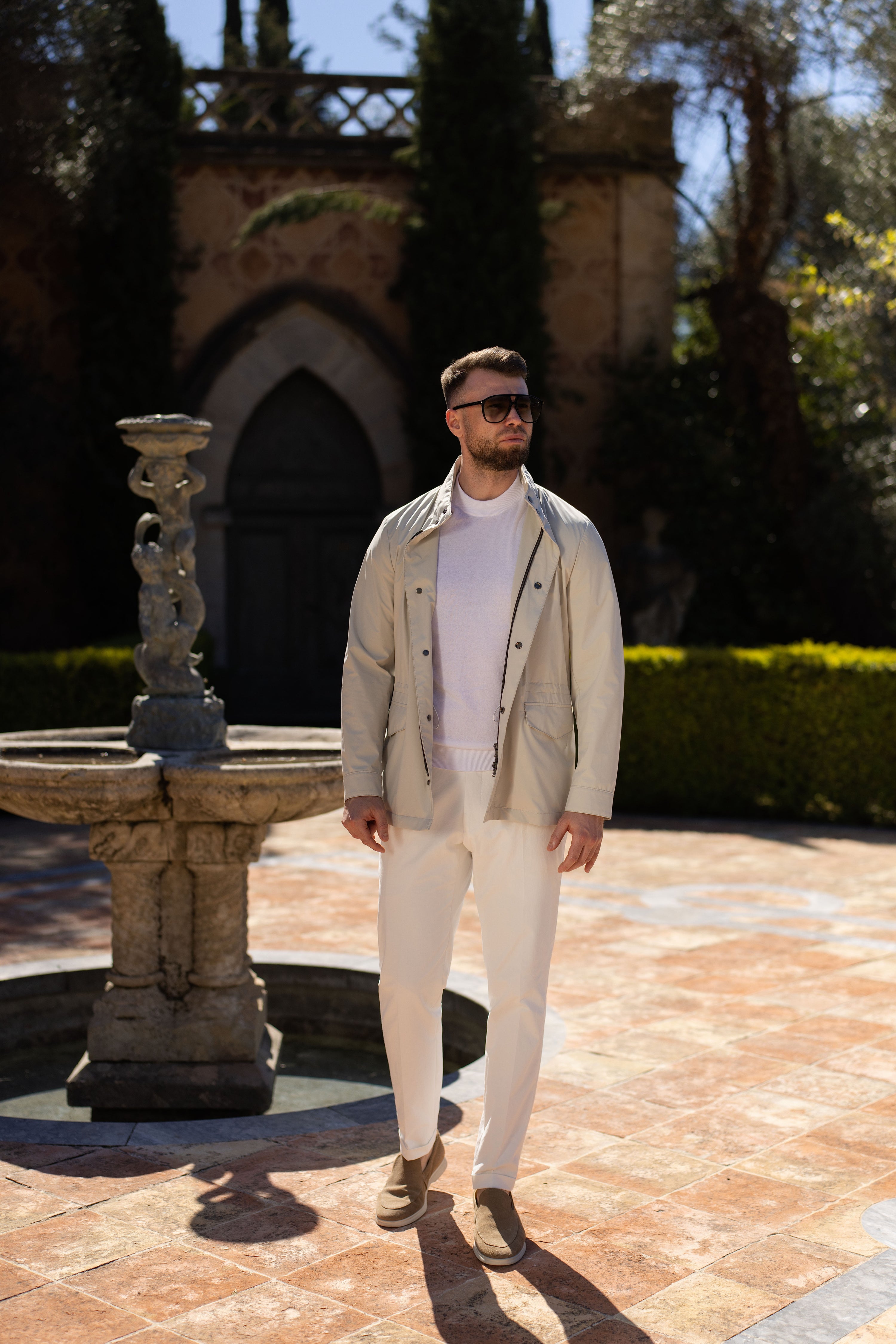 Light-coloured blouson in a complete look, in a garden setting with a fountain