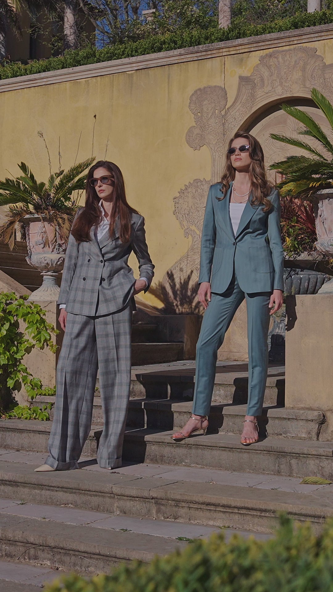 Two women in a grey checked suit and a teal suit on garden steps
