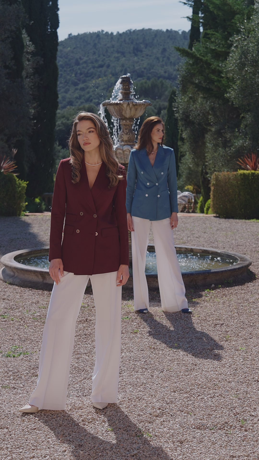 Two women in burgundy and blue jackets with white trousers in a garden