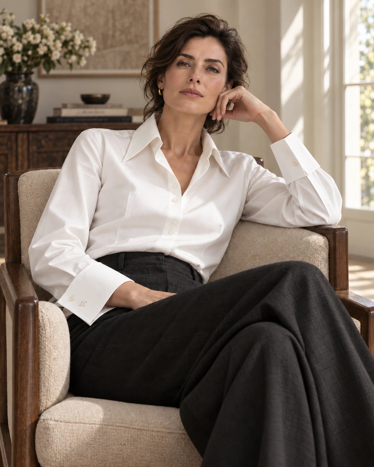 Woman in an ivory shirt with a generous collar and shaped cuffs, relaxed in an armchair, looking at the camera, with charcoal trousers.